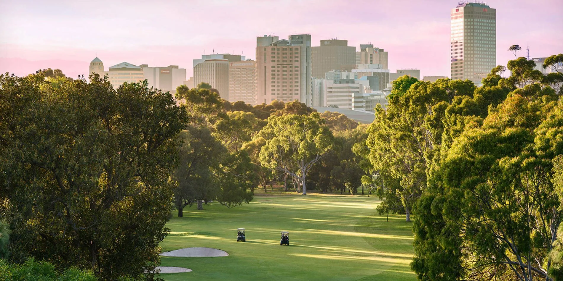 North Adelaide Golf Course at dusk — lush fairways framed by parkland trees with the Adelaide skyline beyond